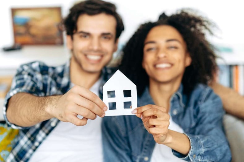 New home owners Joyful multiethnic couple holding paper house, sitting on a sofa in their new modern apartment or house